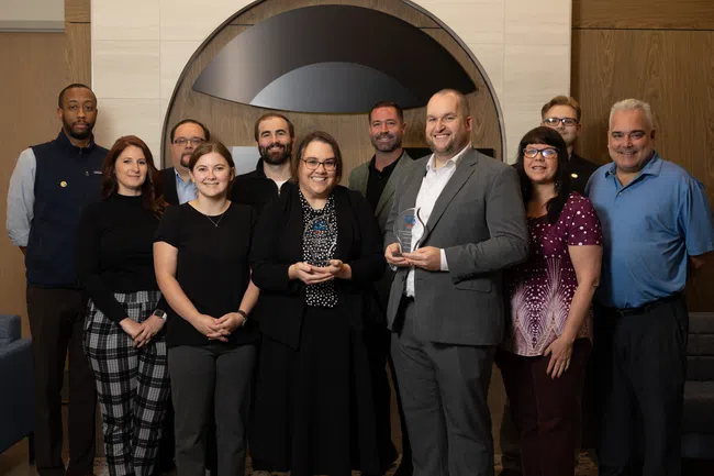 Group of eleven diverse professionals smiling and holding awards in front of a modern office backdrop.