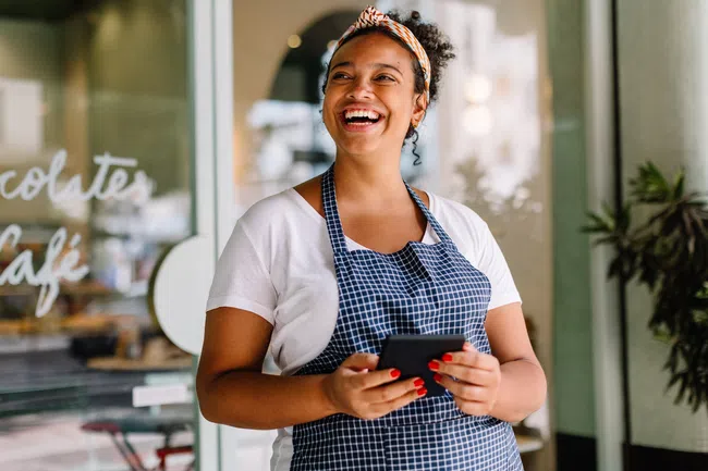 Smiling barista wearing apron and headband holds mobile phone inside modern cafe with glass windows.