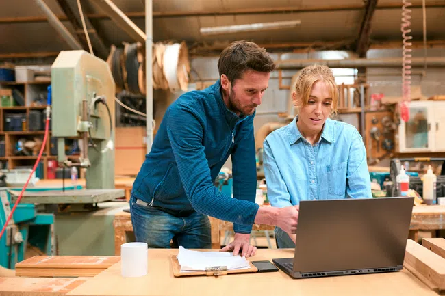 Two coworkers collaborate on a laptop in a busy woodworking workshop filled with tools and materials.
