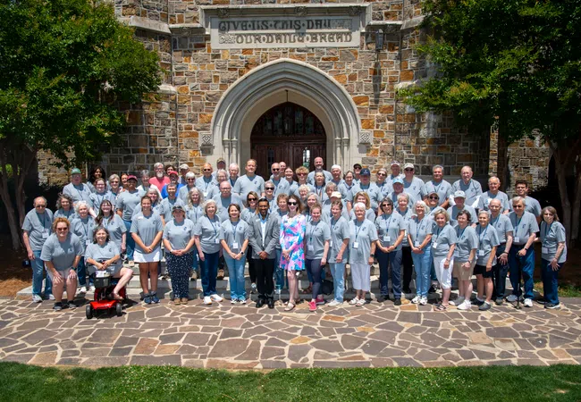Large group of people wearing gray shirts posing in front of a stone building with an arched entrance. Well-lit outdoor scene.