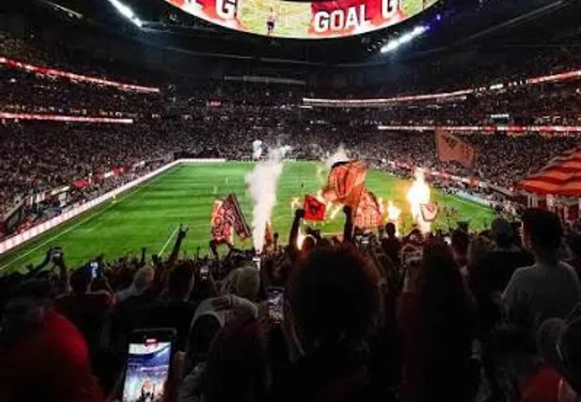 Excited soccer fans celebrating a goal in a packed stadium under a large digital scoreboard at night.