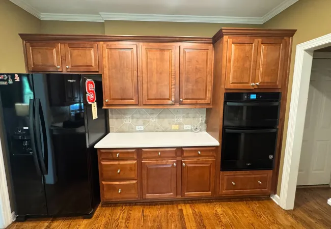 Kitchen with wooden cabinets, black Whirlpool refrigerator, built-in double oven, and white countertop on hardwood floor.
