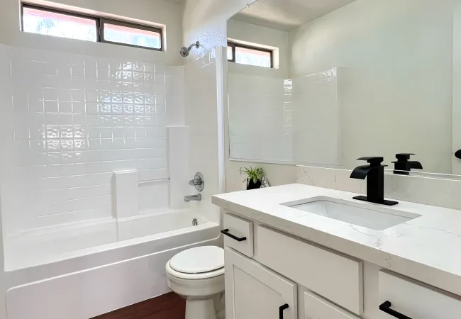 Modern white bathroom with a bathtub, black faucet, large mirror, and dark wood flooring under natural light.