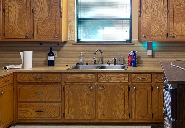Vintage kitchen with wooden cabinets, double stainless steel sink, window with blinds, and countertop essentials.