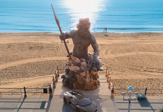 Statue of Poseidon holding a trident on a beachfront promenade with a sports car parked nearby