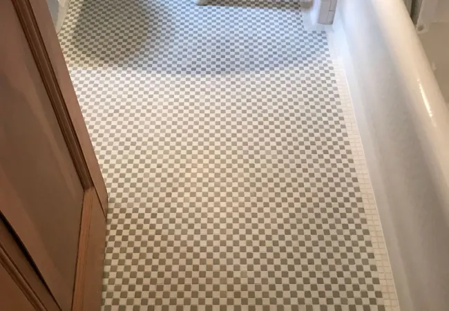 Bathroom floor with vintage gray and white checkered tiles beside a wooden door and white fixtures.