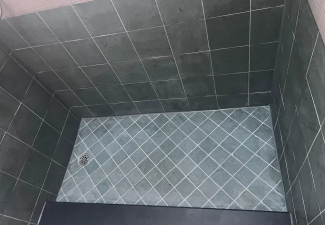 After photo of a small bathroom shower with dark gray wall tiles and lighter gray floor tiles with diagonal grout lines.