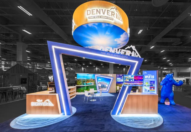 Modern Denver tourism booth with illuminated signage, digital displays, blue bear statue, and seating area.