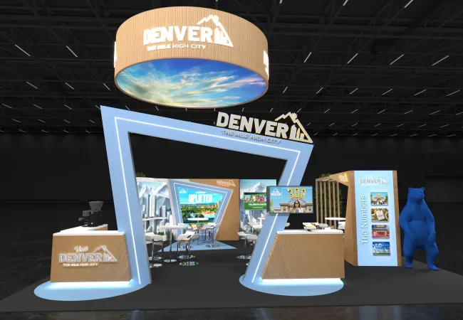 Modern Denver tourism booth with large signage, digital displays, and a blue bear statue in a dark venue.