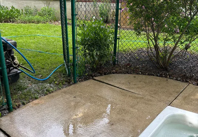 Wet patio with concrete slabs next to a white tub and green chain link fence near a blooming lilac bush.