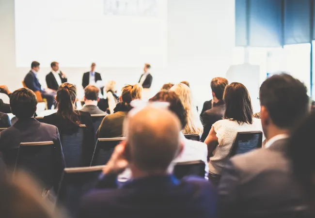 Audience attending a business panel discussion in a bright conference room with speakers on stage.