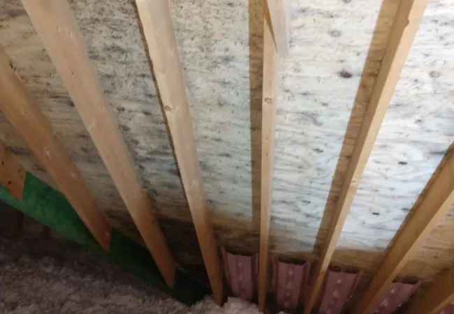 Attic space showing wooden rafters, plywood roof decking, and loose-fill insulation on attic floor.