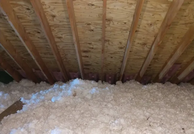 Attic space with wooden beams and loose-fill fiberglass insulation spread over the floor.