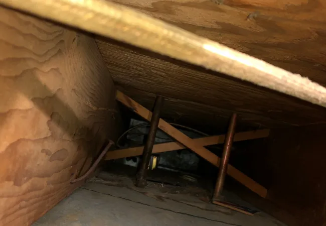 View inside a wooden crawl space with support beams, metal pipes, and plywood walls and floor.