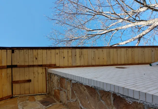 Wooden fence and gate next to stone wall and tiled surface under clear blue sky with leafless tree branches