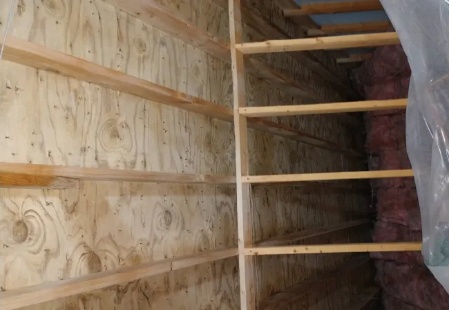 Interior view of attic with wooden beams, plywood walls, and pink fiberglass insulation on the right side.