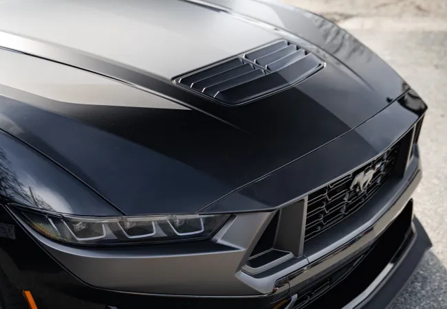 Close-up of a sleek black Ford Mustang front with aerodynamic hood scoop and aggressive headlights on pavement.