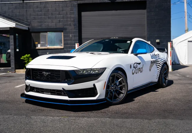 White Ford Mustang with black and blue accents parked in front of a dark building under clear sky.