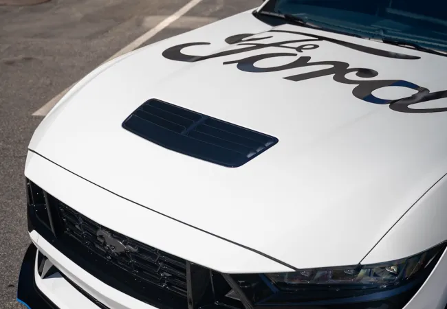 White Ford Mustang with black grille, blue side mirror, and custom hood parked in a lot on sunny day.