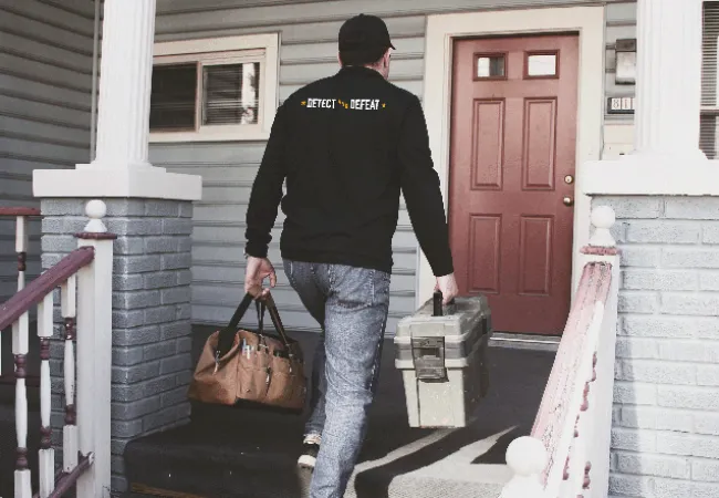 Man carrying tool bags walking up porch steps toward a red front door of a house with gray siding.