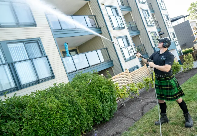 Man wearing a kilt pressure washing an apartment balcony and siding on a sunny day.