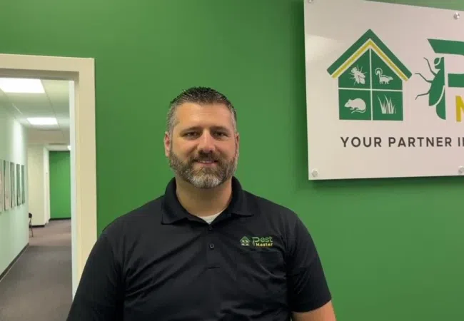 Man in black Pest Master polo shirt sitting by green wall with Pest Master pest control sign.