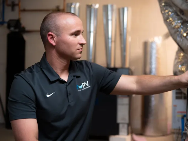 Technician in PV uniform adjusting HVAC system components in a utility room with metal ducts and pipes.