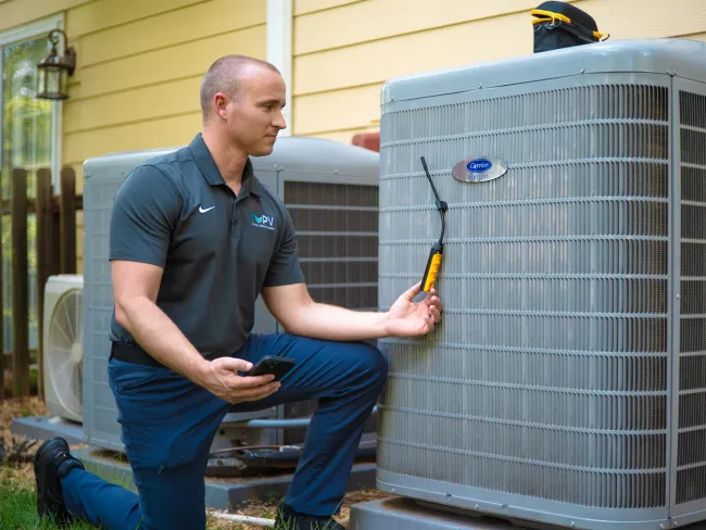 Technician inspecting an outdoor Carrier air conditioning unit using a diagnostic tool and smartphone in daylight.