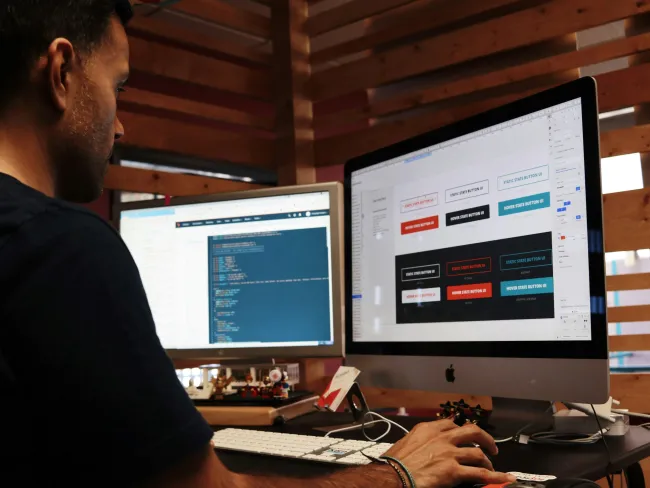 Man coding and designing buttons on dual monitors in a wooden office setting.