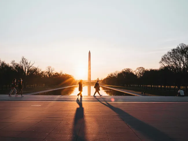 Silhouettes of people walking by the Washington Monument at sunset, reflecting on the water.
