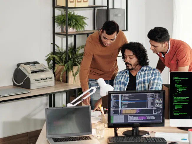 Three developers collaborate on a project in a modern office with multiple screens displaying code and data.