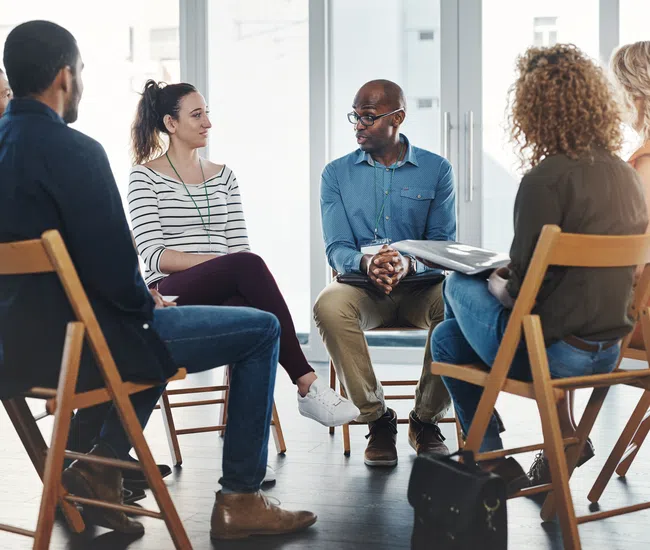 Diverse group of people seated in a circle discussing in a bright room during a support group session.