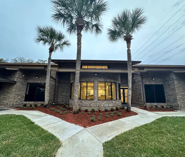 Modern brick building with large windows, palm trees, concrete paths, and landscaped garden beds at dusk.