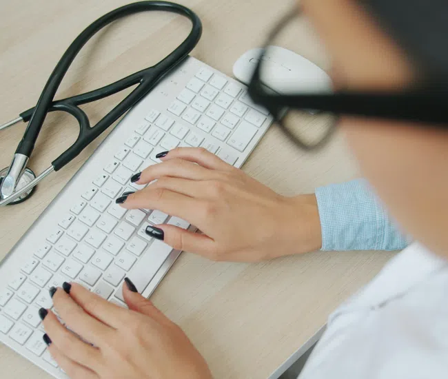 Doctor typing on a white keyboard with a stethoscope placed beside it on the desk