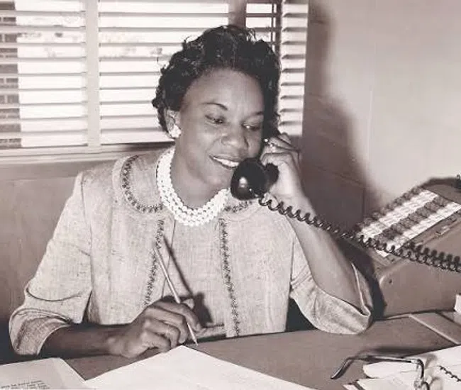 Black and white photo of a woman on a telephone at her office desk with papers and an adding machine