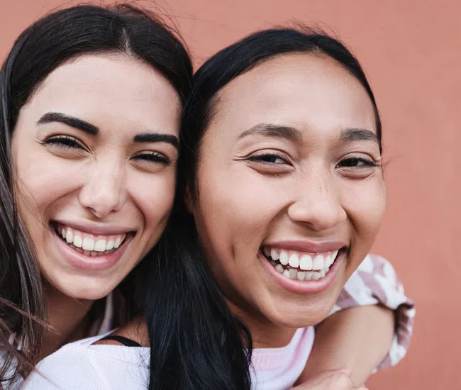 Two happy young women smiling closely together against a peach background, showing friendship and joy.
