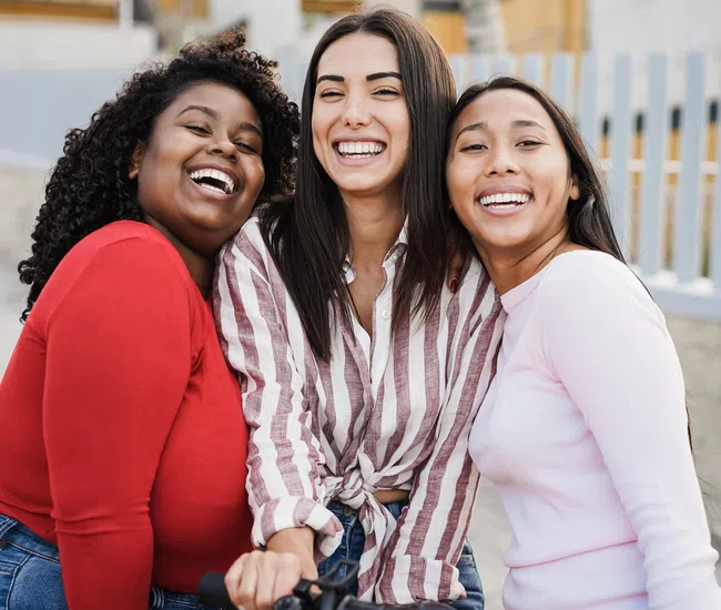 Three diverse young women smiling and hugging outdoors near a fence on a sunny day