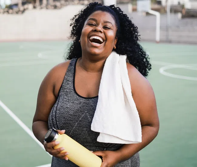 Smiling woman in sportswear holding water bottle with towel on her shoulder on outdoor sports court.