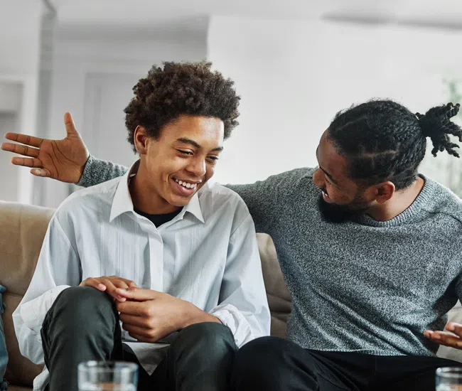 Two African American men smiling and chatting together on a couch in a bright living room.