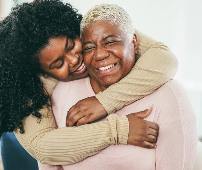 Young woman with curly hair hugging and smiling with an older woman with short blonde hair indoors.