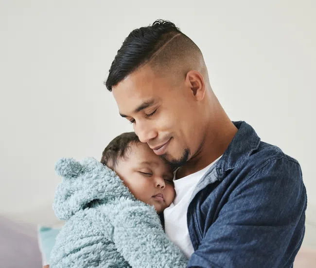 Father tenderly holding and cuddling his sleeping baby wrapped in a soft blue hoodie indoors