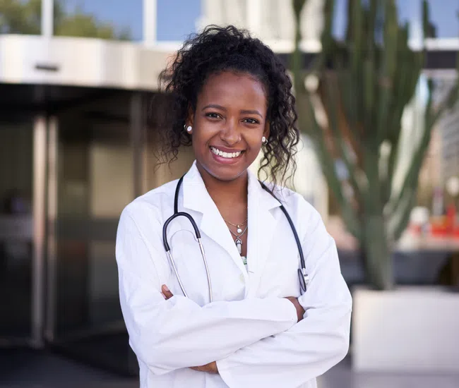 Smiling female doctor with stethoscope in white coat standing outdoors with arms crossed.
