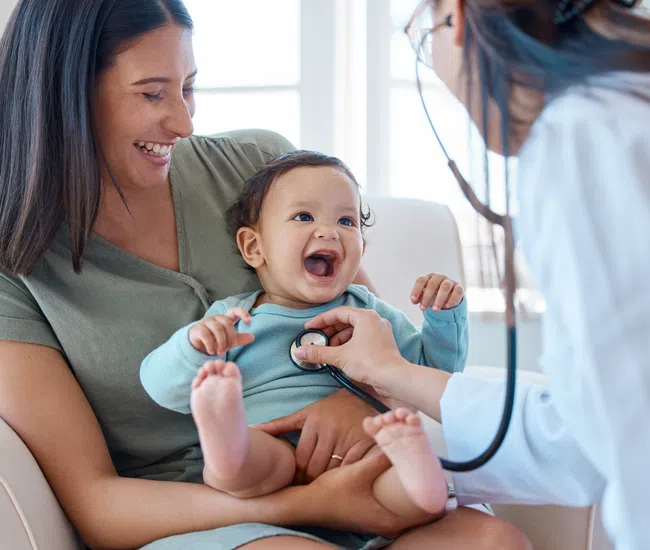 Happy baby being examined by a doctor with a stethoscope while sitting on his mother's lap in a bright clinic.
