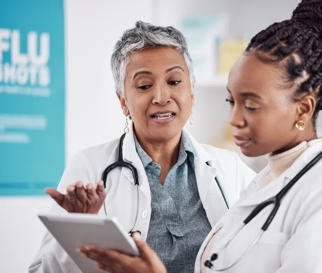 Two female doctors discussing patient information on a tablet in a medical office with flu shots sign