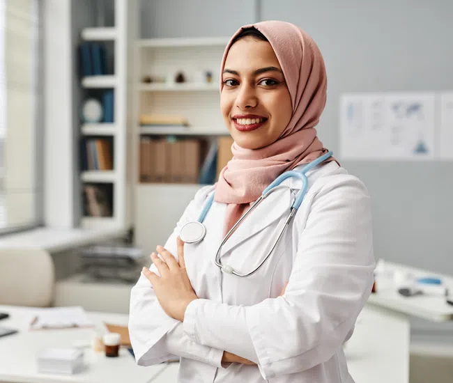 Confident female doctor wearing a pink hijab and white coat with stethoscope in a modern medical office.