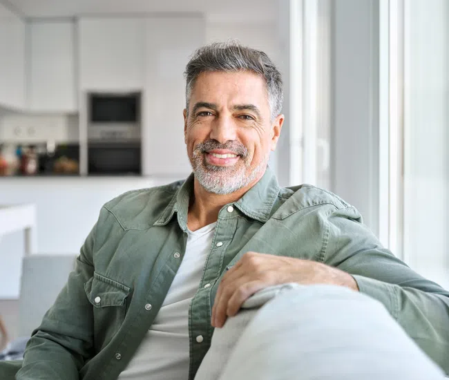 Middle-aged man with gray hair and beard smiling while sitting on a couch in a modern living room.