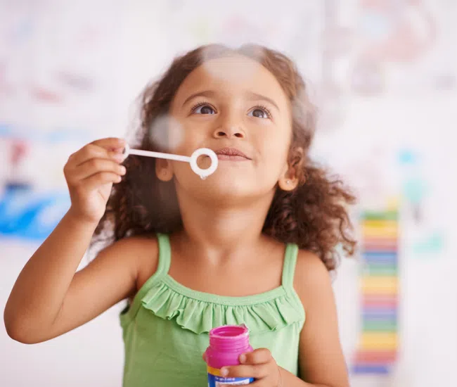 Young girl in green dress blowing bubbles indoors with joyful expression and colorful background.