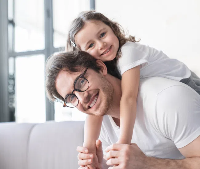 Smiling father with glasses giving piggyback ride to happy young daughter in bright living room.