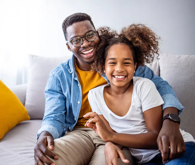 Smiling father and daughter sitting on a gray couch with mustard pillow, enjoying quality time together.