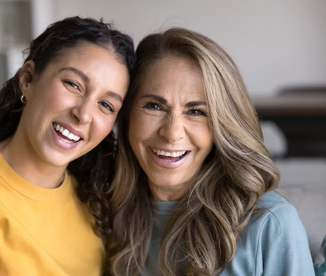 Smiling middle-aged woman with long blonde hair and young woman with curly dark hair in yellow shirt hugging.